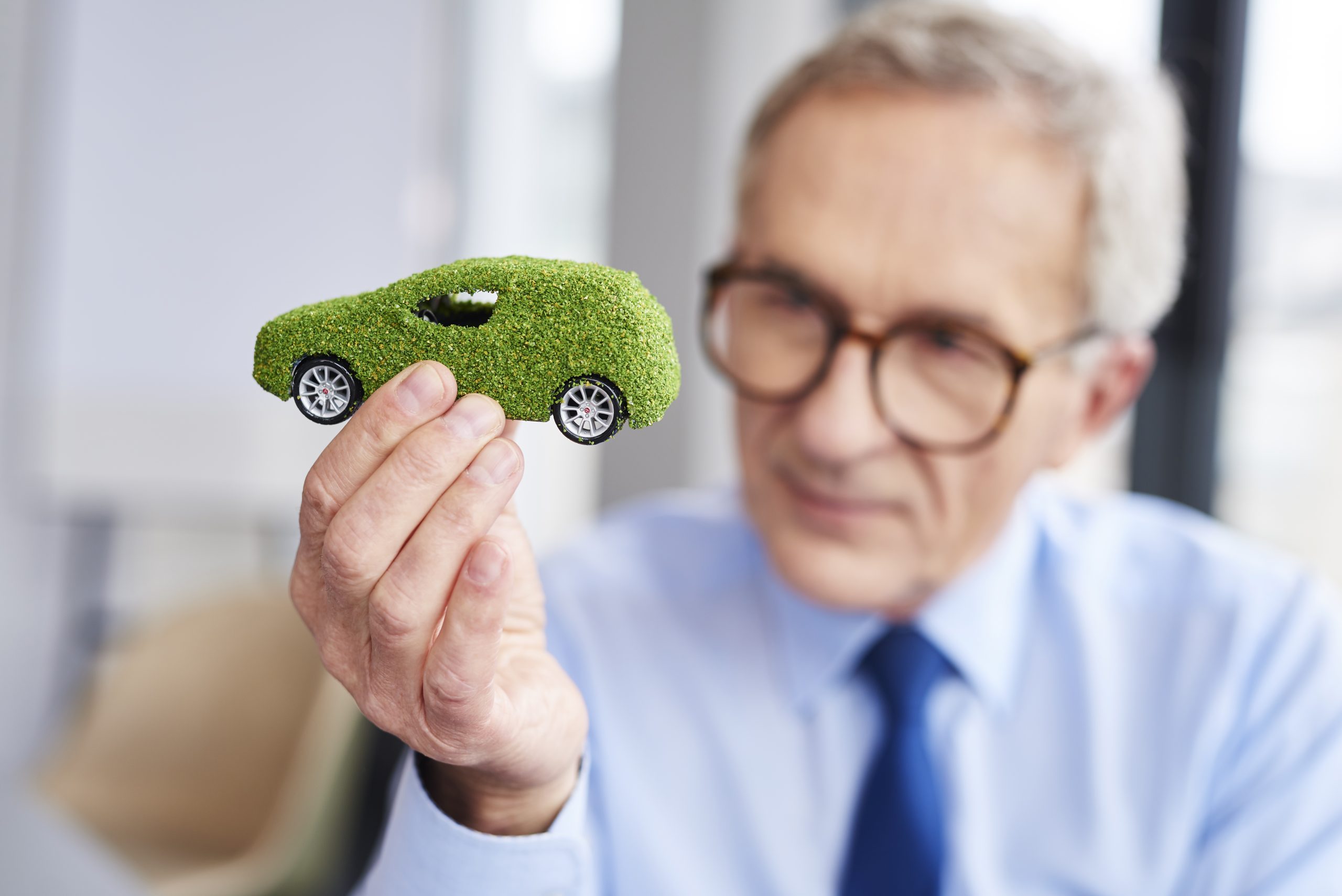 Älterer Mann mit Brille und Krawatte hält ein kleines Modellauto in der Hand, das mit grünem Gras überzogen ist