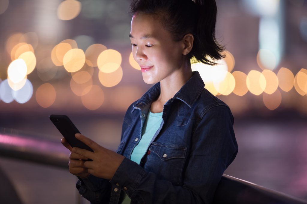Asiatische Frau blickt auf der Straße bei Nacht auf ihr Handy und lächelt