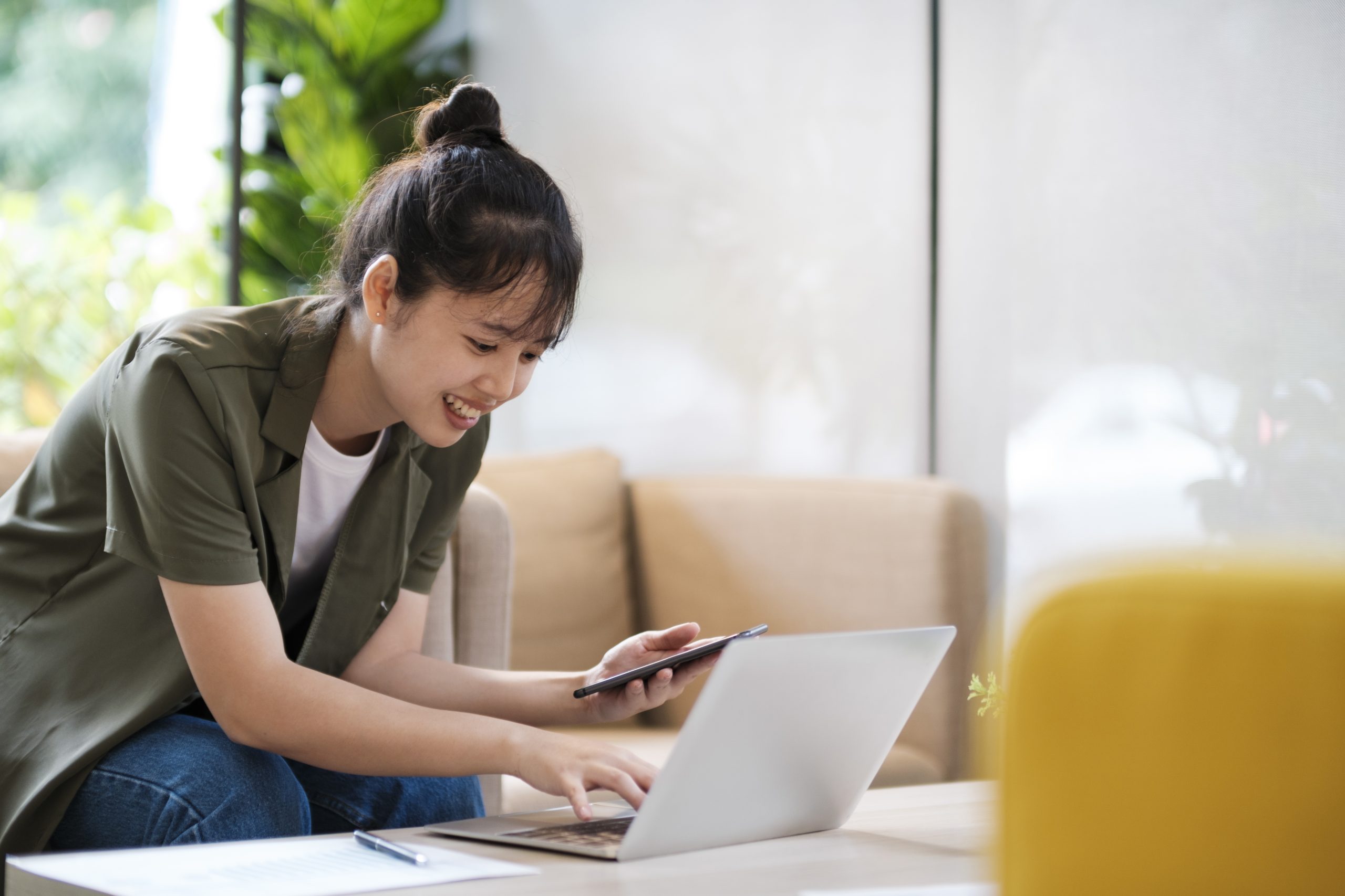 Junge asiatische Frau hält Handy in der Hand und schaut auf Laptop