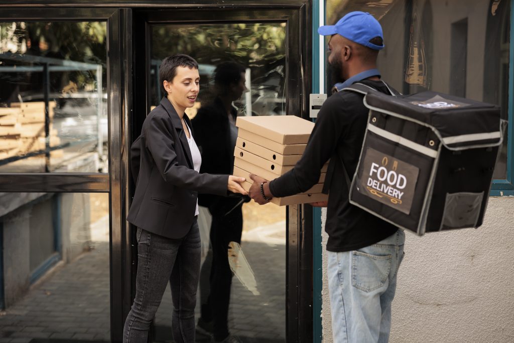 Ein Mann mit blauer Kappe und schwarzem Pulver hat eine Kühltasche in schwarz am Rücken und überreicht an einer Haustür acht Pizzaschachteln an eine Dame in schwarzem Hosenanzug