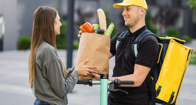 Mann mit gelbem Rucksack überreicht einer Frau in Jeans und Pullover eine Papiertüte mit Lebensmitteln auf der Straße