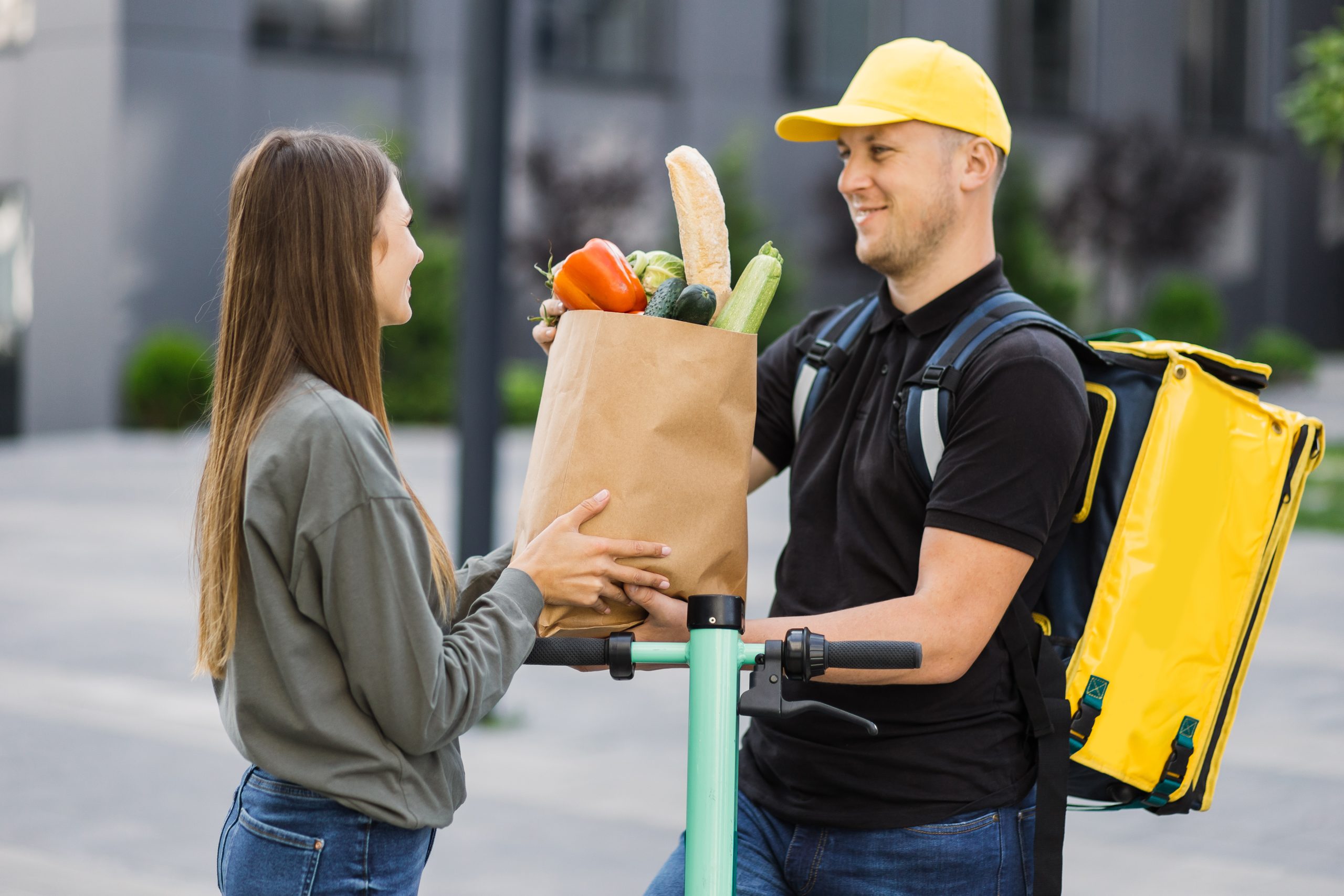 Mann mit gelbem Rucksack überreicht einer Frau in Jeans und Pullover eine Papiertüte mit Lebensmitteln auf der Straße