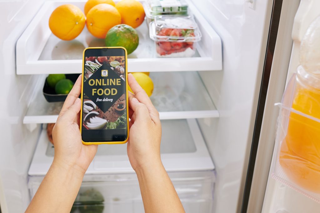 Kühlschrank in dem sich Obst und Gemüse befinden davor zwei Hände die ein Handy halten und am Display Online Food free delivery zu sehen