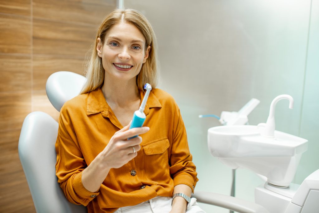 Junge hübsche Damen mit einer orangen Bluse hält eine elektrische Zahnbürste in der Hand und sitzt auf einem weißen Sessel in einer Zahnarztpraxis