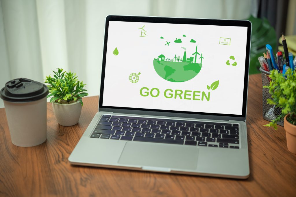 Ein aufgeschlagenes Notebook das auf dem Bildschirm GO GREEN und einige Symoble in grün sowie eine halbe Erdkugel mit einem Haus, Windrädern und Fabriken in grün zeigt