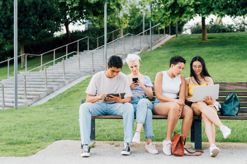 drei junge Menschen sitzen in Sommerkleidung auf einer Parkbank ud schauen auf ihre Notebooks sowie Handys