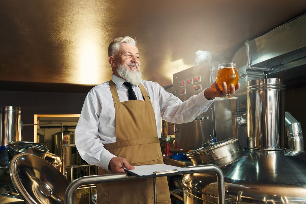 Mann mit weißen Haaren und Bart hält ein Glas mit frisch gebrautem Bier in der Hand