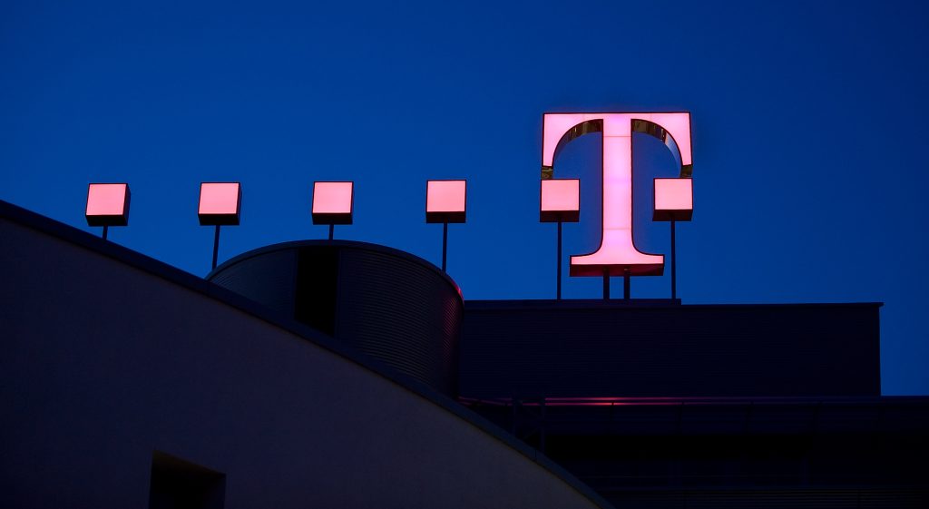 Logo der Deutschen Telekom in Form eines riesigen T in pink mit pinken Punkten auf einem Gebäude montiert. Nachtansicht mit dunkelblauem Himmel
