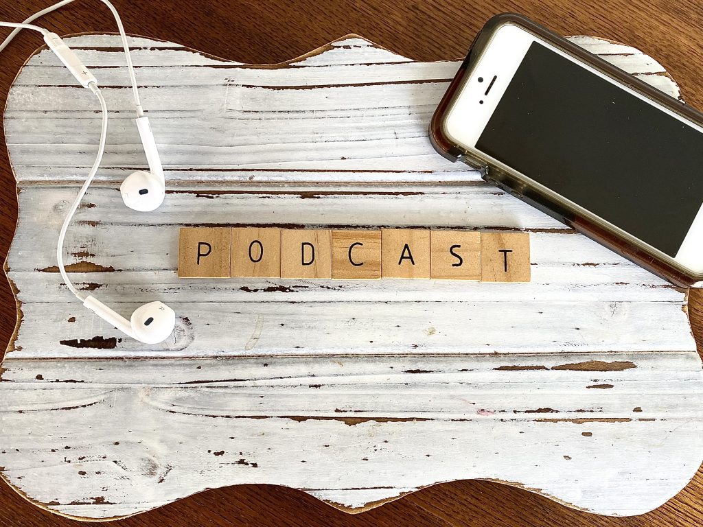 Brauner Tisch auf dem ein Schild liegt indem Podcast auf braunen Klötzen geschrieben steht. Darüber ein Headset sowie ein Handy