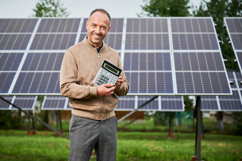 Junger Mann steht vor einer Solaranlage und hält einen großen Taschenrechner in Händen