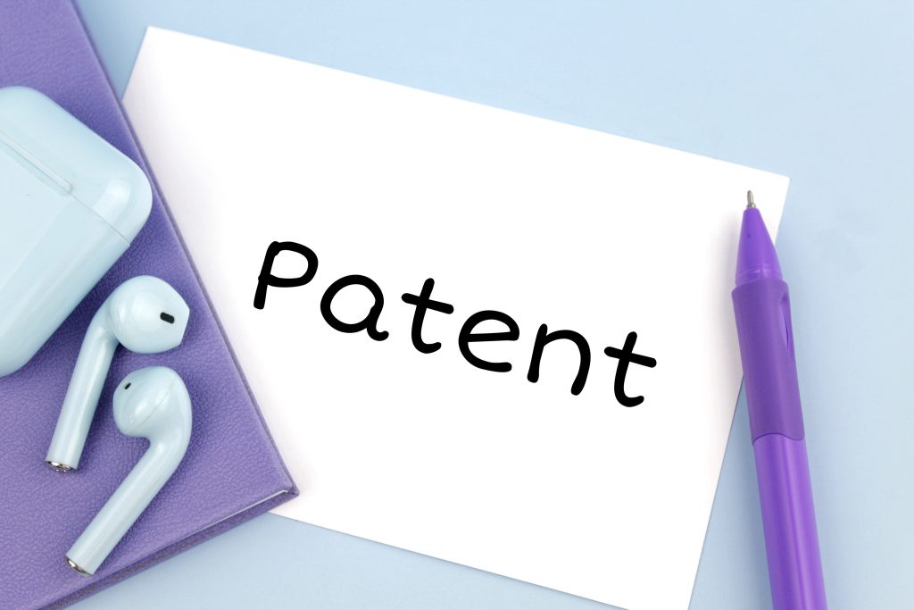 Weißer Zettel mit dem Wort Patent. Rechts davon ein Kugelschreiber in violett. Links davon zwei Ohrenstöpsel auf einem violetten Notizbuch