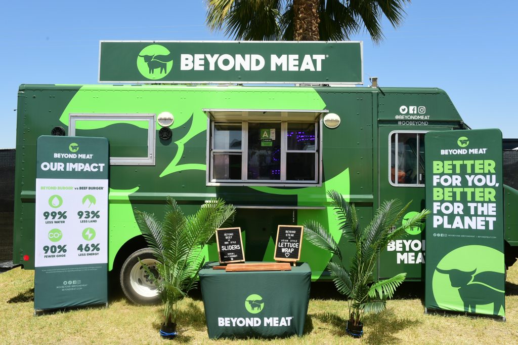 Verkaufsstand von Beyond Meat in Grün unter einer Palme. Davor zwei Roll-ups und ein grüner Tisch mit Holzbrett und Logo von Beyond Meat