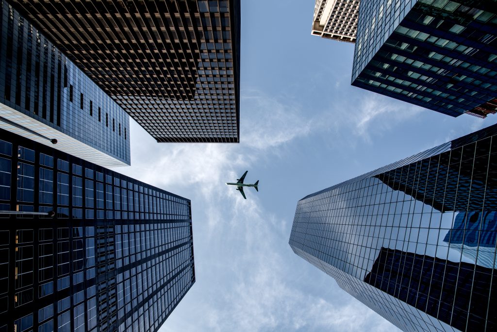 Blick von unten fotografiert auf fünf Hochhäuser über die ein Flugzeug fliegt