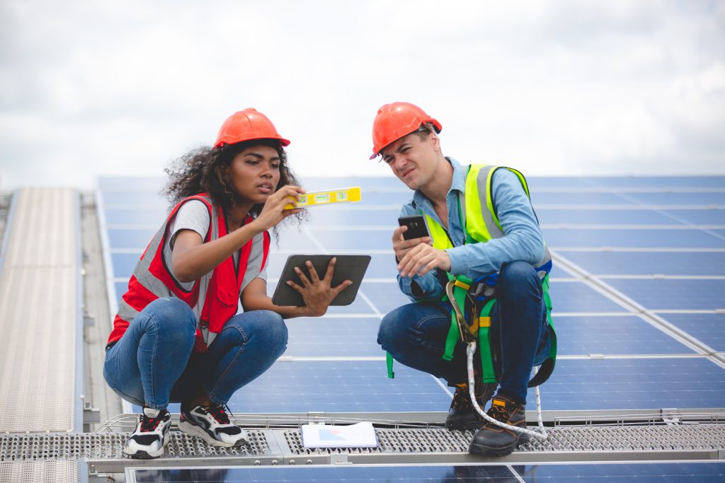 Frau und Mann montieren eine Solaranlage. Sie hält ein gelbes Messgerät und ein Tablet, beide hocken und er hält ein Handy in der Hand