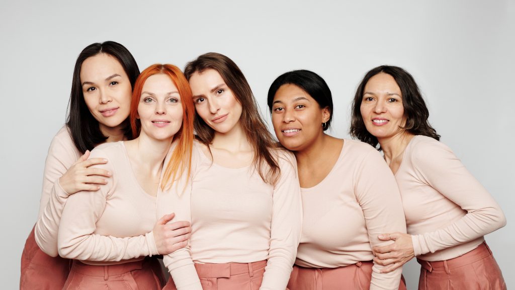 5 Frauen mit hellen T-Shirts und dunklen Haaren sowie rosa Hosen blicken in die Kamera